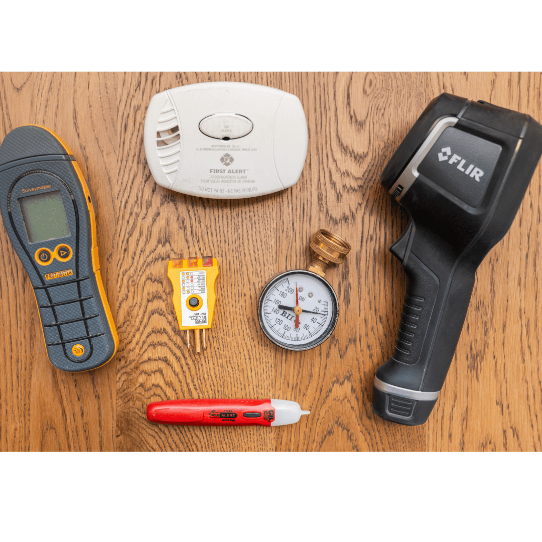 Various diagnostic tools including a moisture meter, smoke detector, infrared thermometer, electrical outlet tester, pressure gauge, voltage detector, and infrared camera on a wooden surface.