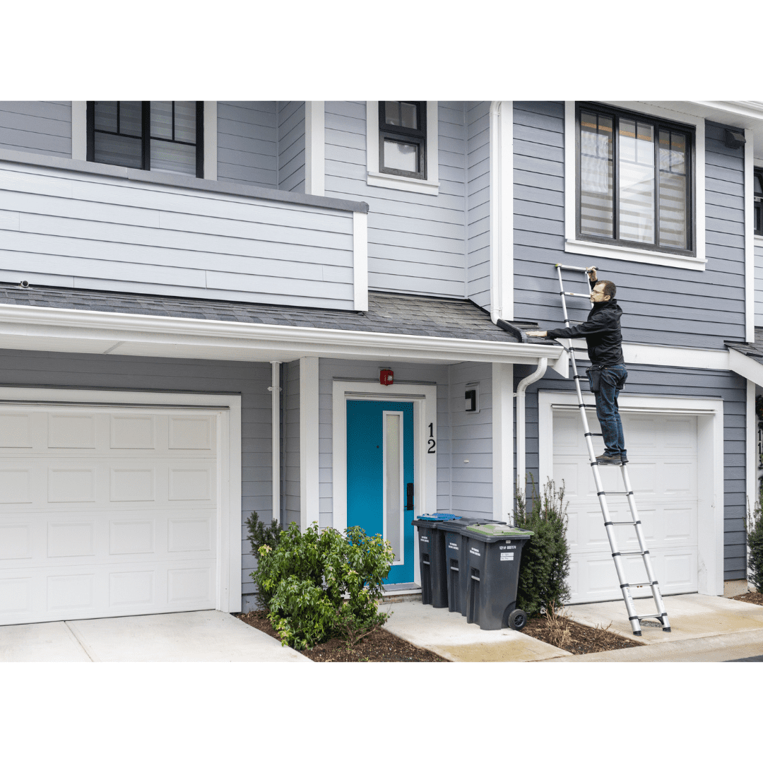 A person stands on a ladder cleaning the gutters of a two-story house with blue siding, white trim, and a blue front door. Three trash bins are located near the entrance. The house number is 12.