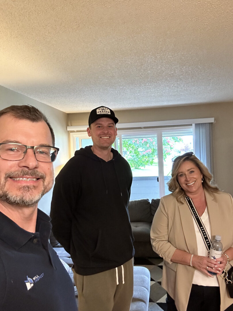 Three people after home inspection pose for a photo inside a living room, smiling. One person is holding a disposable cup, another person is wearing a cap, and the third person is wearing a blazer and holding a bottle.