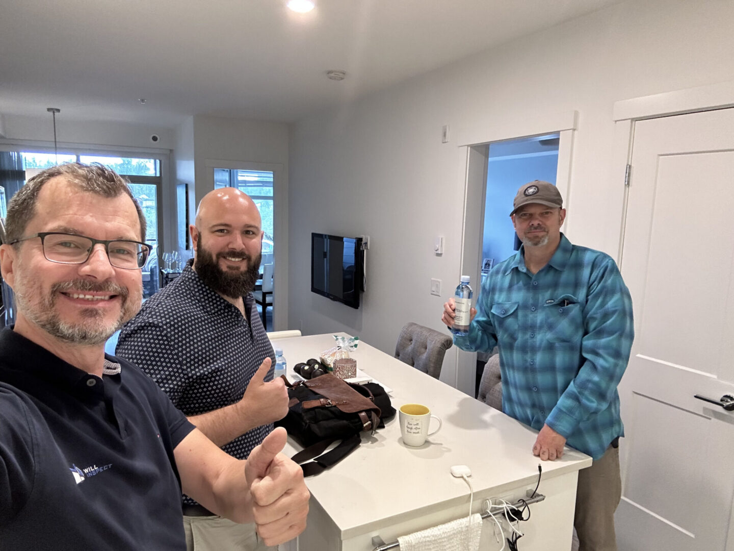 Three men are standing in a kitchen, smiling at the camera. One gives a thumbs-up, another holds a drink, and the table has various items like a mug and electronic devices.