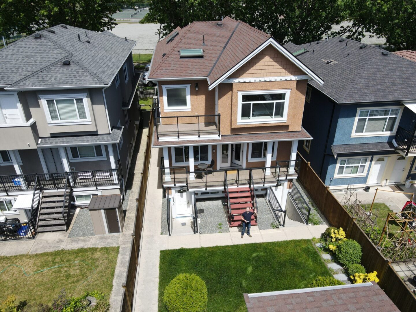 Aerial view of a two-story house with a well-maintained lawn in the backyard. A person is sitting on a chair in the backyard. Surrounding houses have similar architectural styles.